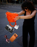 Bolso Sailor daidai · qué cabe dentro · modelo sujetando el bolso naranja junto a cámara, auriculares, móvil y gafas de sol · Fūwa